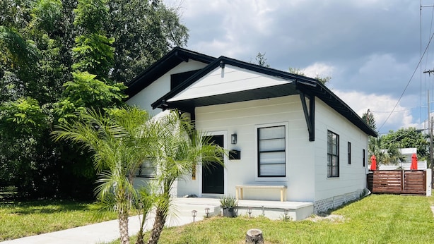 Una casa de un solo piso con fachada blanca y detalles en negro, un pequeño porche con un banco y un césped bien cuidado.