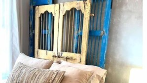 Una cama con colcha y almohadas color beige, una mesita de noche de madera con una lámpara, y una pared con paneles azul y blanco desgastados.