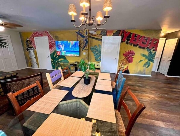A dining area with a large table set for four, surrounded by wooden chairs. There is a chandelier above the table, and the walls are decorated with colorful murals.