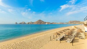 Ein Strand mit Reihen von Liegestühlen und Sonnenschirmen, einem klaren blauen Himmel und einer gebirgigen Kulisse.