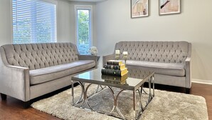 A living room with two grey upholstered sofas, a glass coffee table, and a white shag rug. There are two framed abstract paintings on the wall. - Soléra — A Light-Filled Minimalist Escape Near Beaches & Golf (Innisfil)