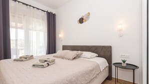 Una cama bien tendida con almohadas y toallas dobladas, una mesita de noche con una planta, y una lámpara fijada en la pared.