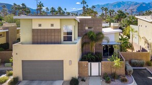 Un edificio residenziale moderno con garage, un piccolo cancello d'ingresso e un giardino anteriore curato con palme e arbusti.