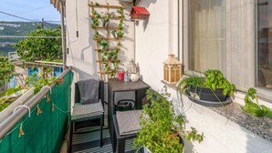 Un balcón con una pequeña mesa, sillas, plantas en macetas, una escalera de madera y un farol rojo.