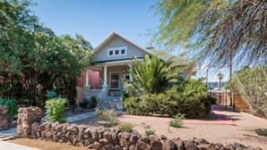 A two-story house with a front porch, stone retaining wall, and surrounding vegetation. - Historic Jostaad Home! (Tucson)
