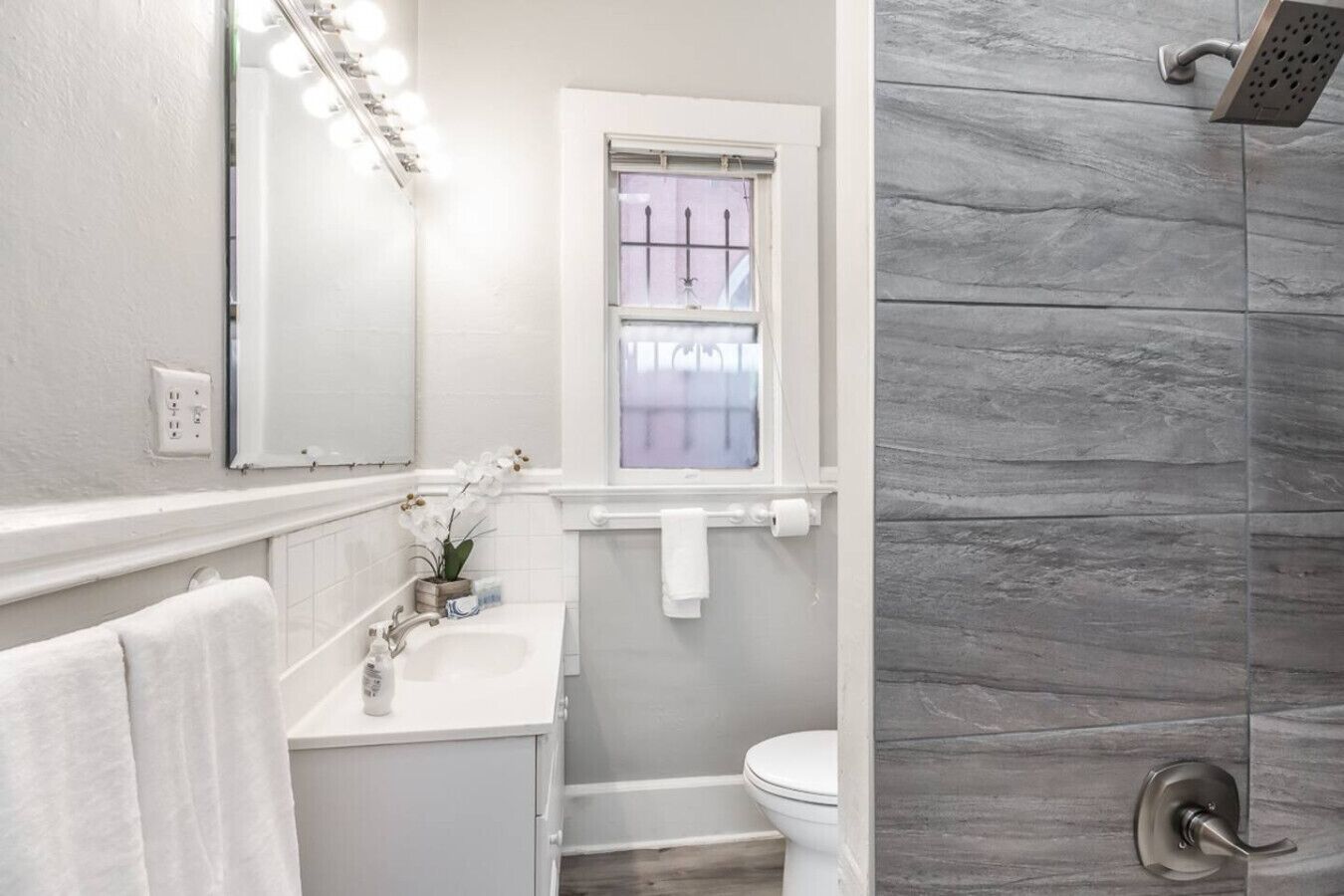 Ein Badezimmer mit weißem Waschbecken, einem beleuchteten Spiegel, einem Fenster, einer Toilette und einer Dusche mit grauen Fliesen.