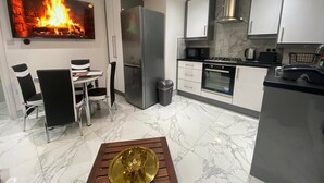 Une cuisine moderne avec des armoires blanches, un réfrigérateur en acier inoxydable, un four à micro-ondes et une cuisinière. On y trouve un coin repas avec une table et des chaises, ainsi qu’un grand tableau représentant un feu au mur.
