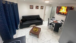 Un salon avec un canapé noir, une table basse agrémentée d’un objet décoratif, un coin repas avec une table et des chaises, et un téléviseur diffusant une scène de feu.