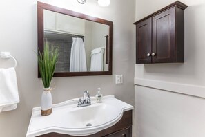 Une salle de bain avec un lavabo blanc, un miroir, un meuble en bois foncé et une plante dans un vase blanc.