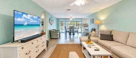 A living room with a television displaying a beach scene, a sofa, a coffee table with fruits, and a ceiling fan.