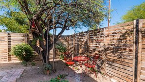 Un espacio al aire libre cercado con un árbol, un jardín pequeño y sillas y una mesa de metal rojo.