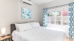 Un dormitorio con una cama, una mesita de noche con lámpara, aire acondicionado instalado en la pared y una ventana con cortinas estampadas.