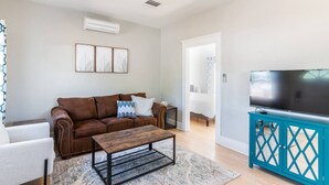 Un salón con un sofá de cuero marrón, una mesa de centro de madera, un mueble azul turquesa con un televisor y una puerta blanca que da a otra habitación.