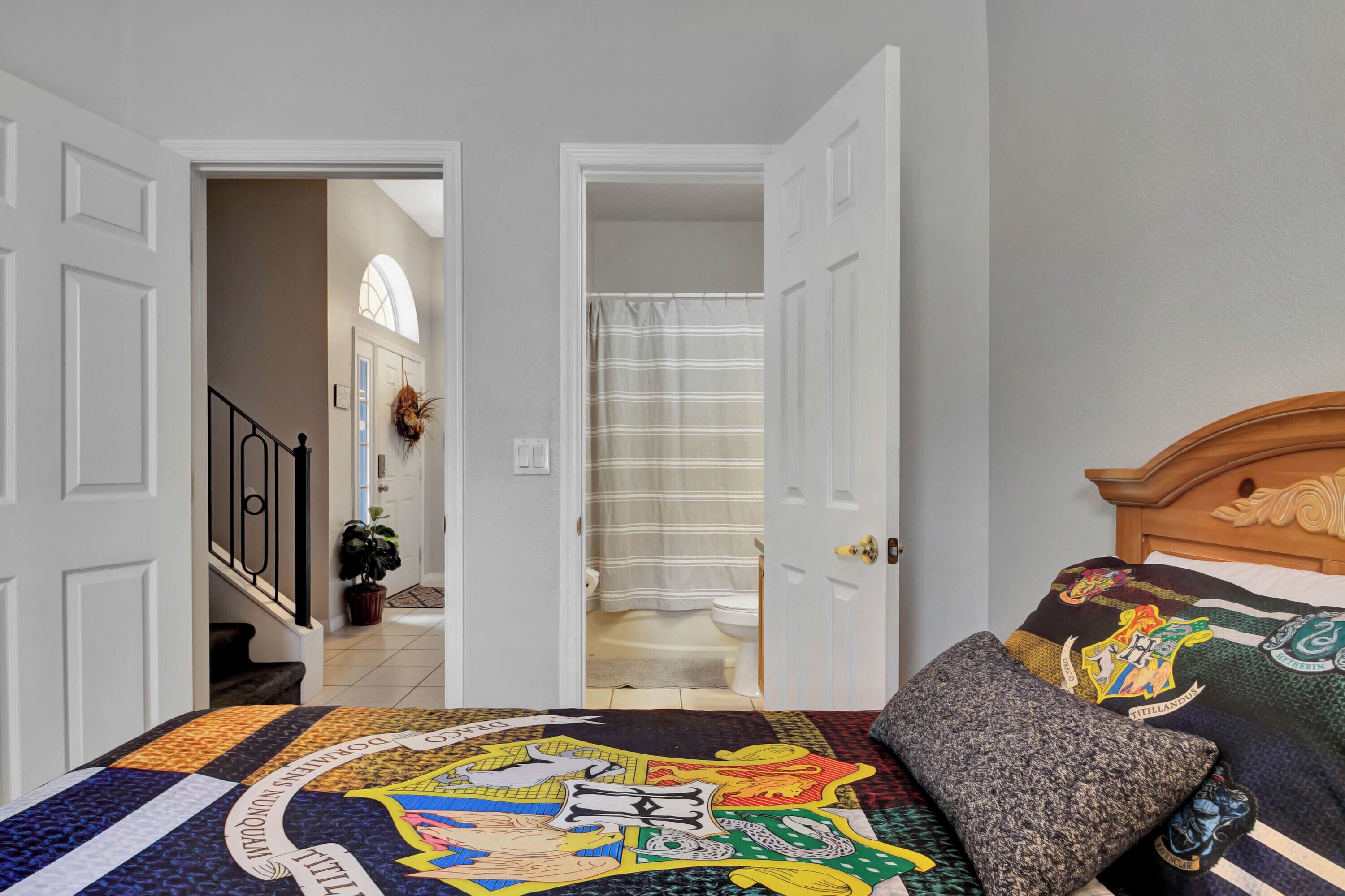 A bedroom with a large bed featuring a Hogwarts crest quilt, a wooden headboard, and a grey pillow. A door leads to a bathroom with a shower curtain.