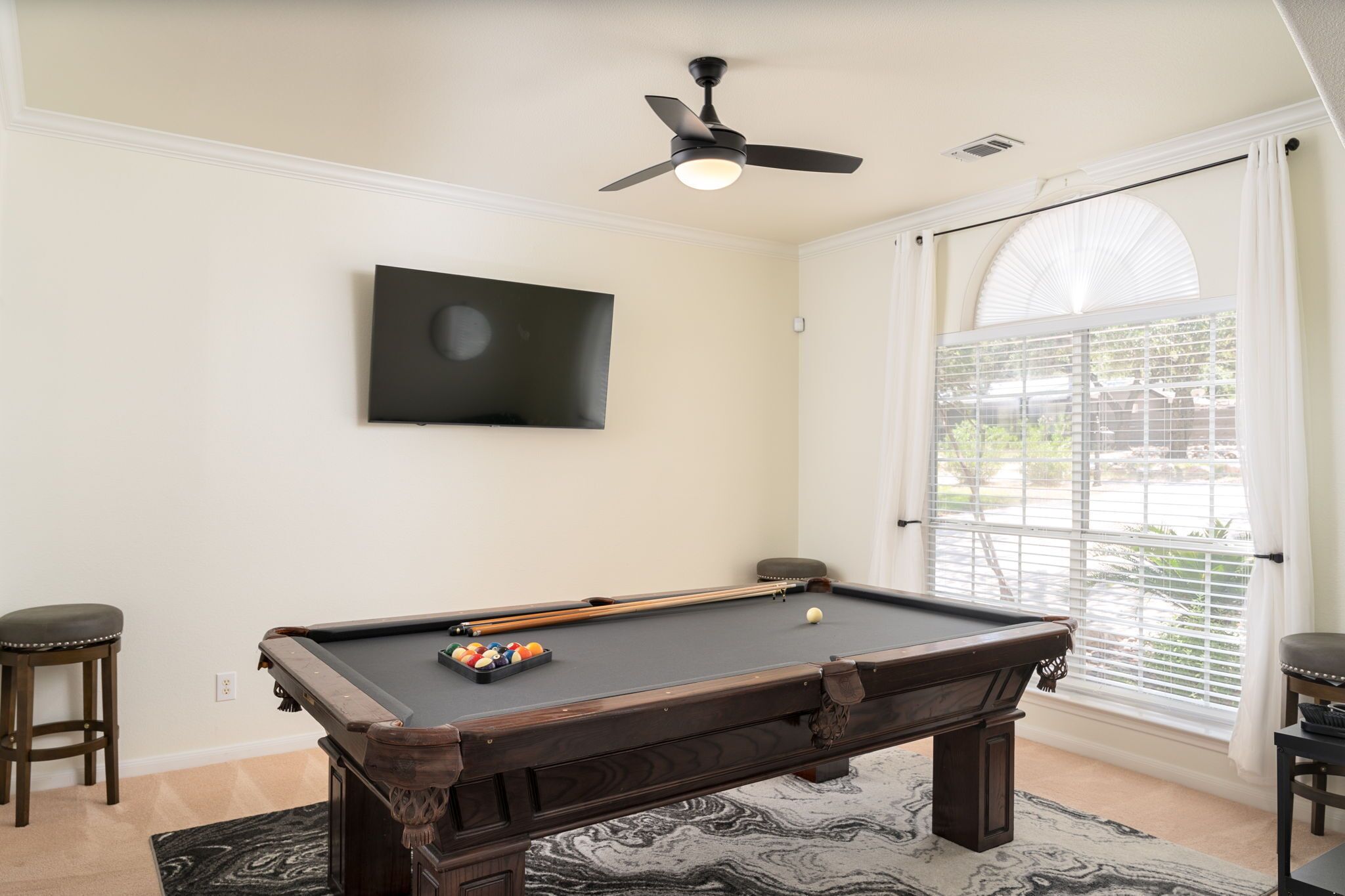 Une table de billard avec des billes, un ventilateur de plafond, un téléviseur fixé au mur et une fenêtre avec des stores.