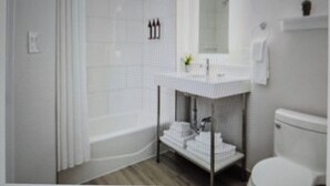 Baño con lavamanos blanco, espejo y inodoro. Se ve una barra de cortina de ducha y un perchero.