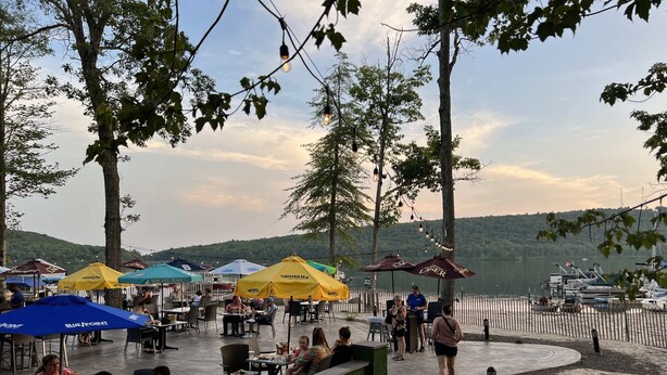 An outdoor dining area with tables and chairs under umbrellas, overlooking a body of water with boats.