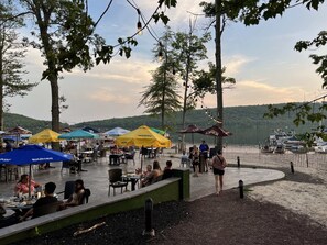 An outdoor dining area with tables and chairs under umbrellas, overlooking a body of water with boats.