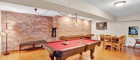 A pool table in a room with brick walls, a dartboard, and a dining table with chairs.