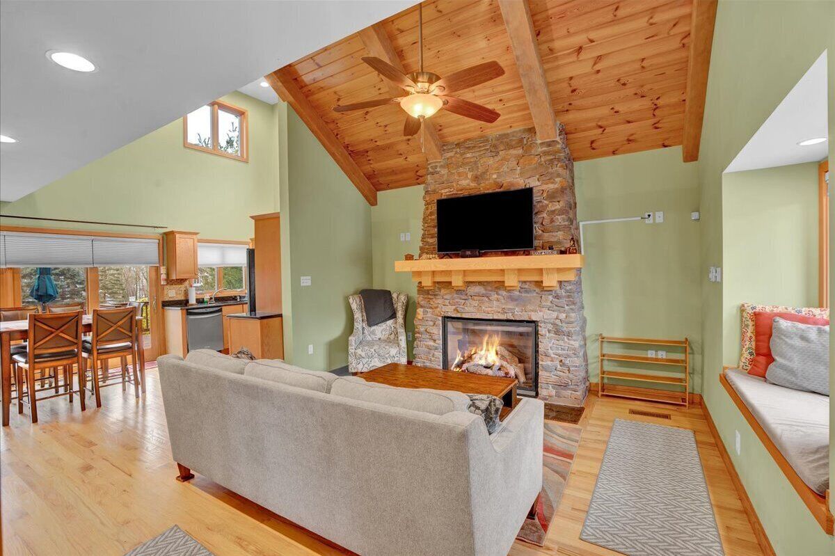 A spacious living room with a stone fireplace, a ceiling fan, and a television mounted above it.