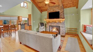 A spacious living room with a stone fireplace, a ceiling fan, and a television mounted above it.