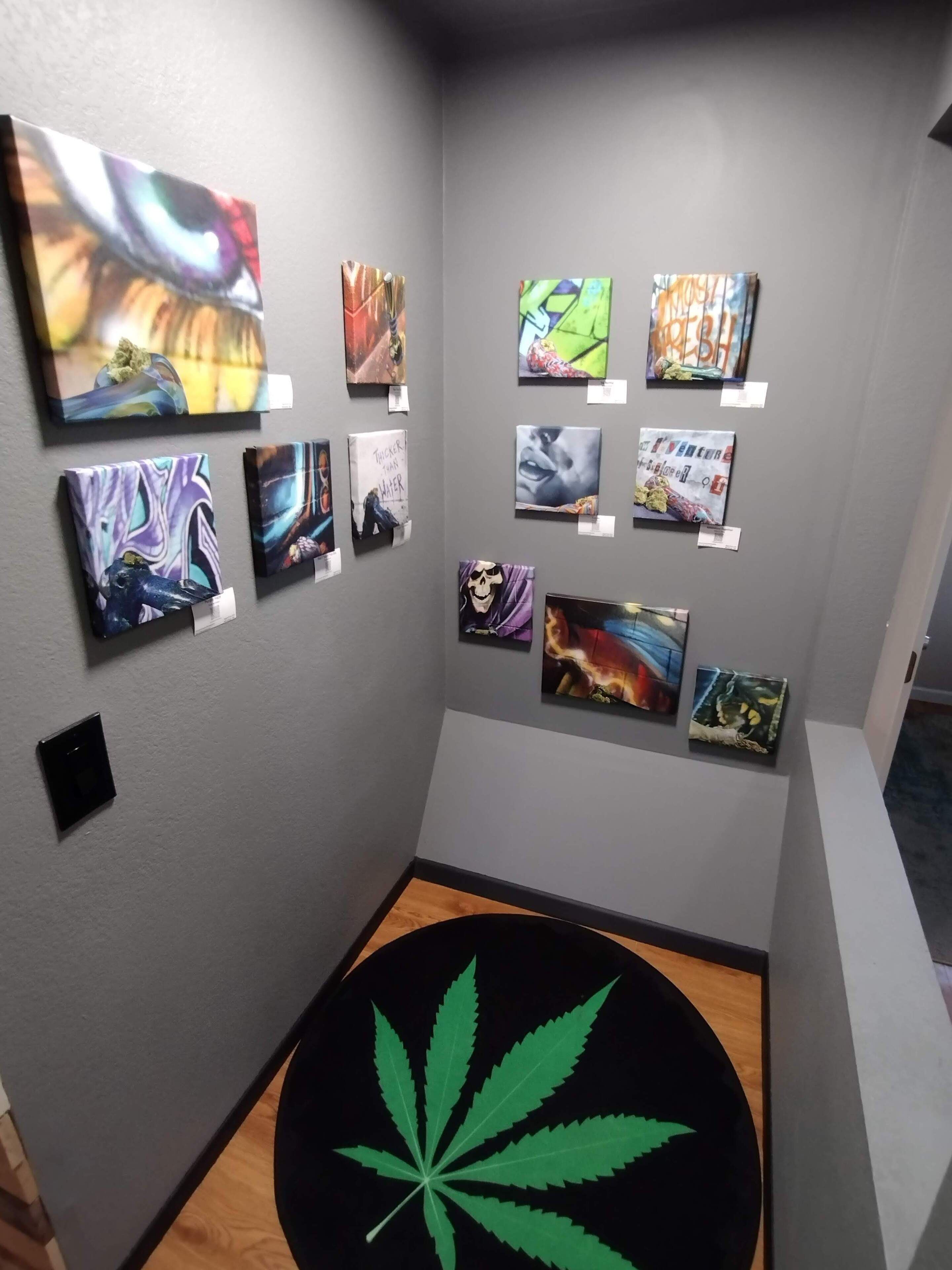 Un mur décoré avec différents tableaux, un objet circulaire noir orné d’un motif de feuille verte, et un interrupteur sur le mur.