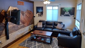 A living room with a wooden wall, a ceiling fan, a black leather sofa, a coffee table with books, and artwork on the walls. - Cannabis Friendly House Minutes From Downtown Denver & Red Rocks (Denver)