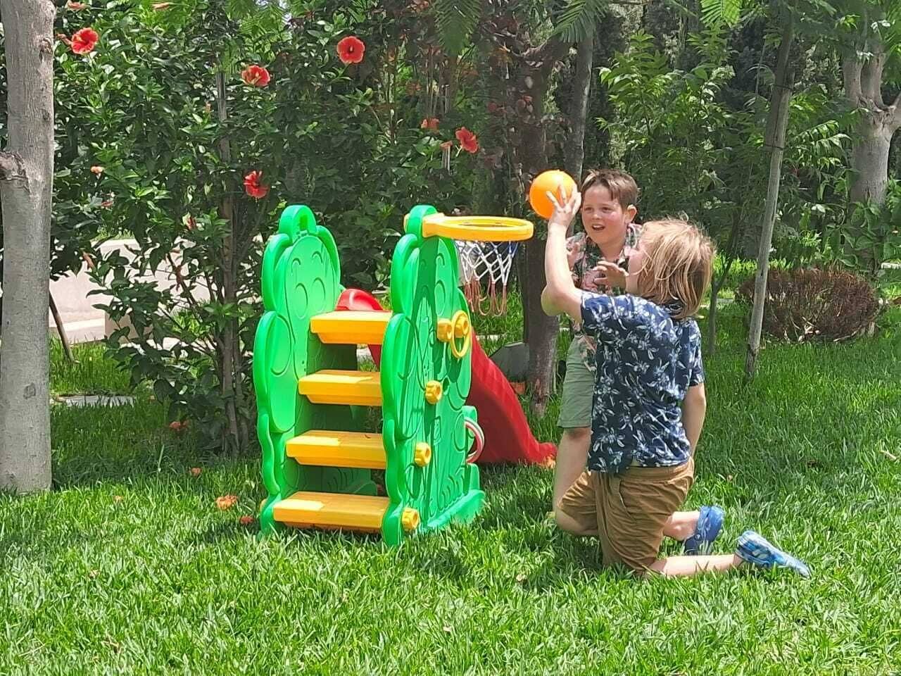 Un enfant joue au basketball sur un terrain vert, avec un panier, entouré d’arbres et d’herbe.