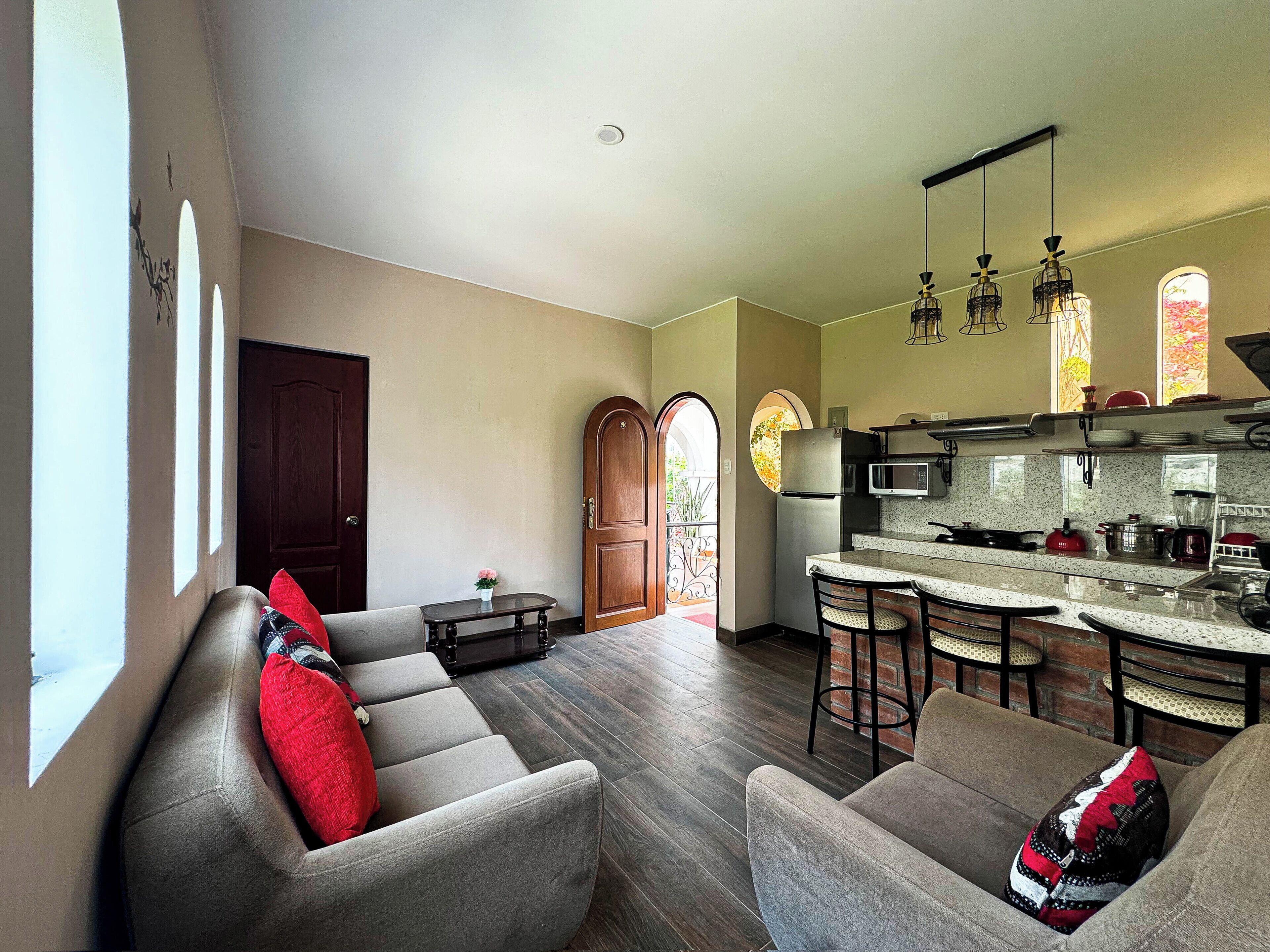 A modern living room with a grey sofa, a small coffee table, and a kitchen area with a refrigerator, microwave, and bar stools.