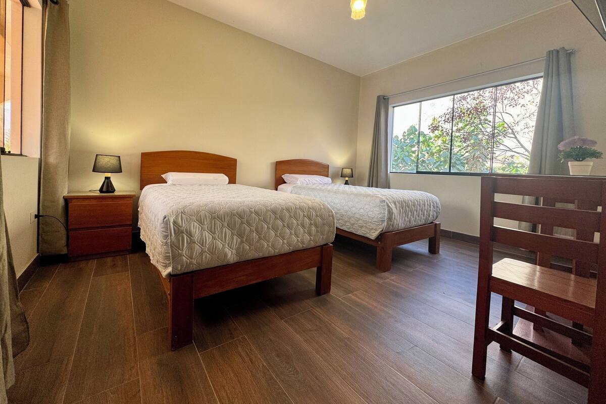 Une chambre à coucher avec deux lits en bois, une table de chevet, une chaise et une fenêtre donnant sur des arbres.