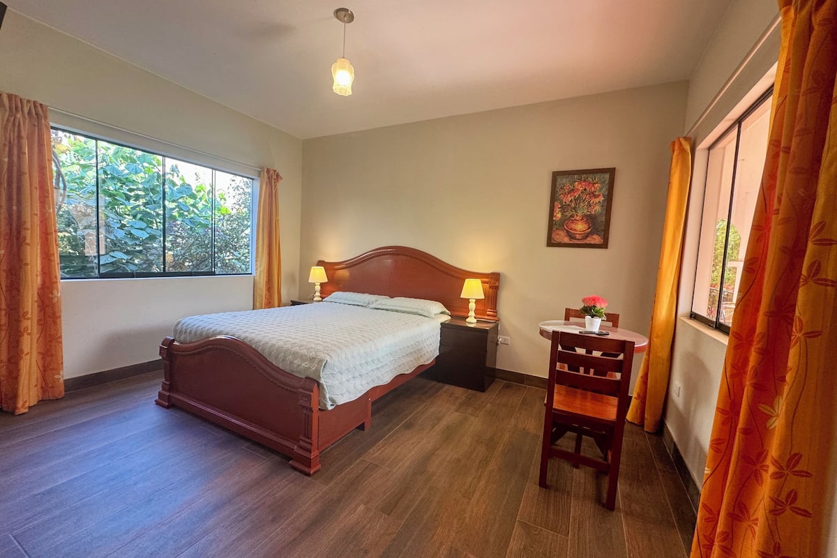 Une chambre à coucher avec un lit en bois, deux lampes de chevet, une petite table avec une chaise et une fenêtre avec des rideaux.