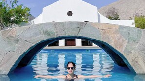 Une piscine surmontée d’un pont en pierre formant une arche, qui mène à un bâtiment blanc doté d’une fenêtre circulaire.
