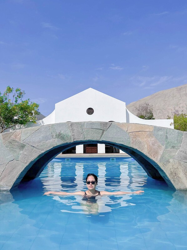 Una piscina con un puente de piedra en arco que conduce a un edificio blanco con una ventana circular.