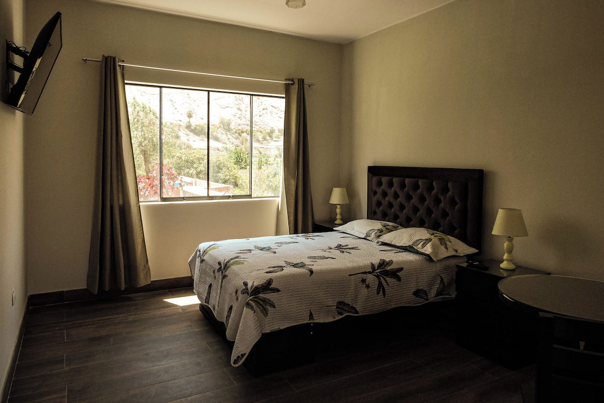 Une chambre à coucher avec un lit, deux lampes de chevet, une table ronde et une fenêtre avec des rideaux.