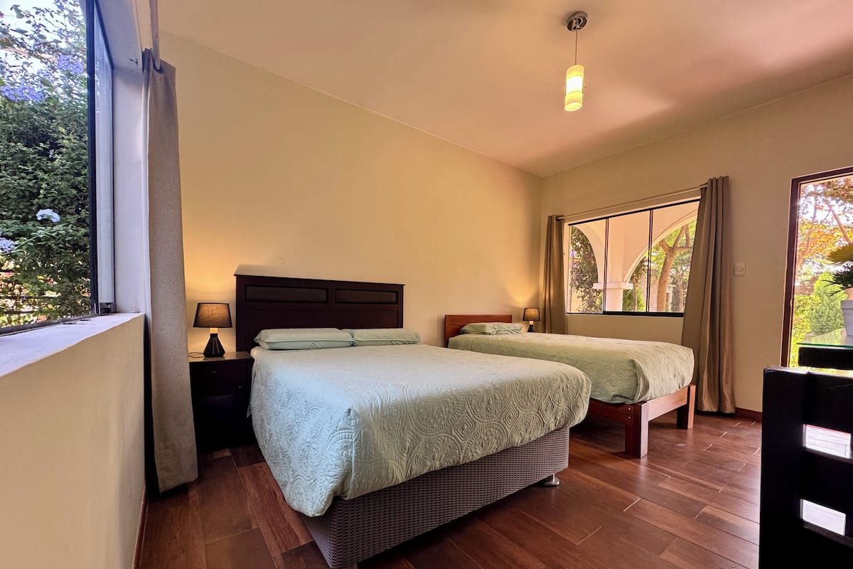 Une chambre d’hôtel avec deux lits, un plancher en bois, une grande fenêtre donnant sur un espace verdoyant et un luminaire suspendu.