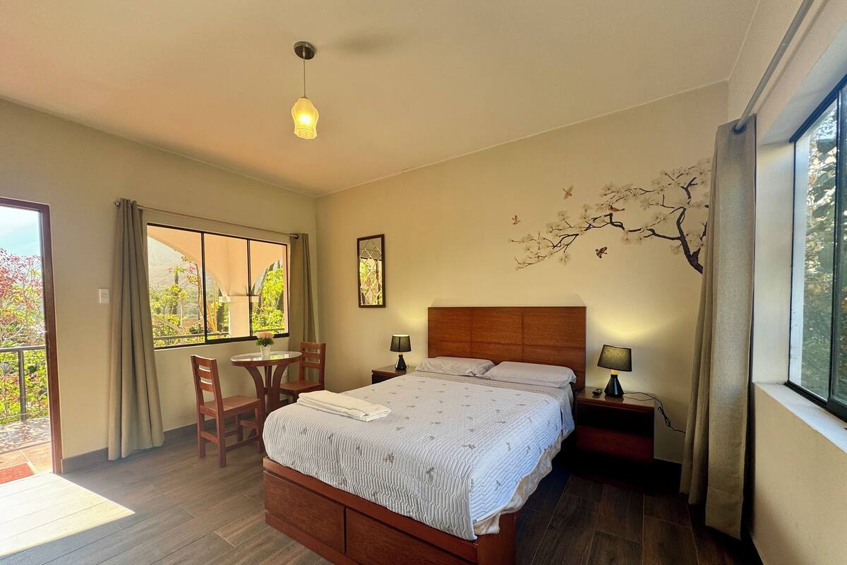 Une chambre à coucher avec un lit en bois, une petite table avec des chaises, une fenêtre avec des rideaux et une lampe fixée au mur.