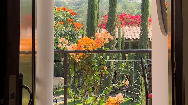 Une vue à travers une porte en arc donnant sur un balcon, avec une chaîne de montagnes en arrière-plan, où l’on aperçoit de grands cyprès et des plantes en fleurs.