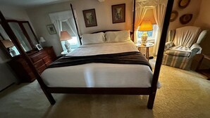 A four-poster bed with a white bedspread, a wooden dresser, a chair, and a small table with a lamp.