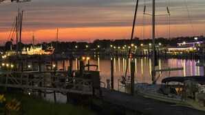 A marina at dusk with boats docked, a wooden pier, and a building with lights in the background.