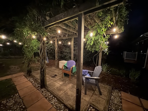Eine Außenpatio mit einer Pergola, Lichterketten, einer überdachten Sitzgelegenheit und einem Garten.