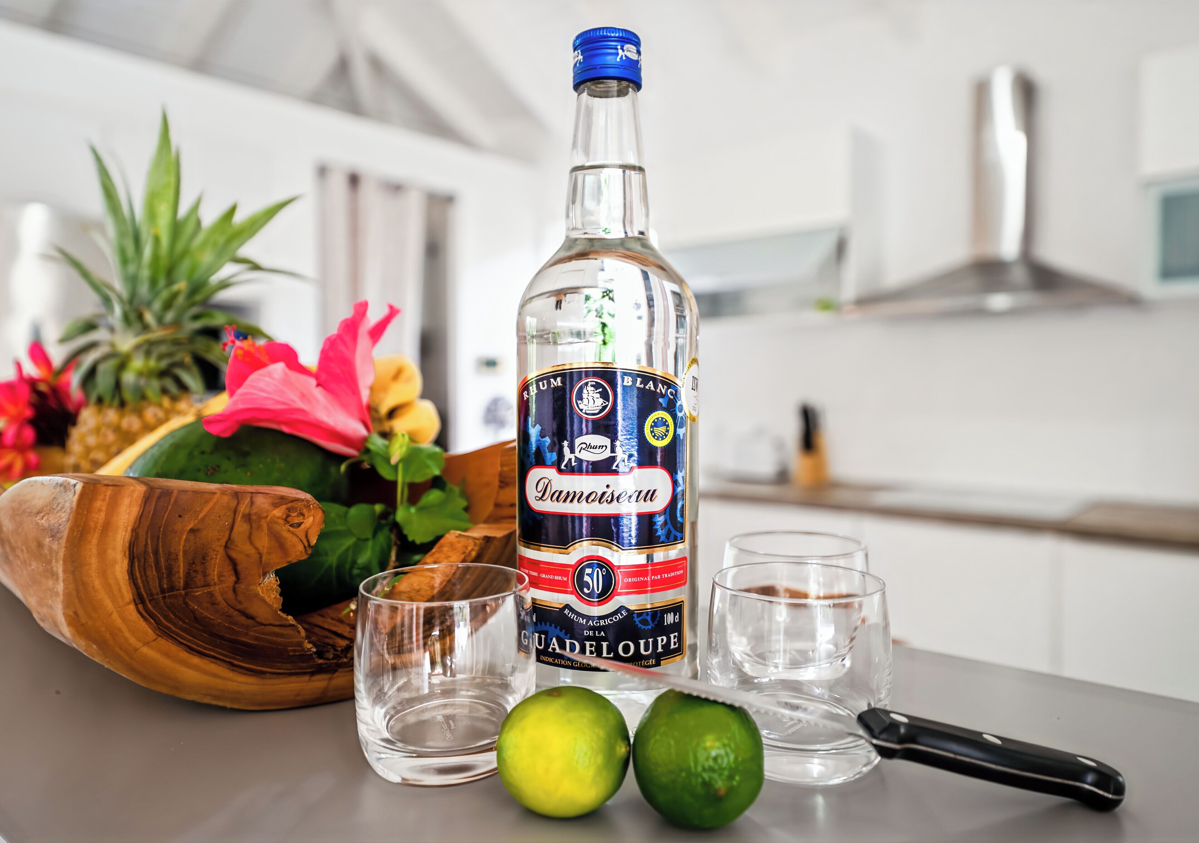 Une bouteille de rhum Damoiseau, deux citrons verts et un saladier en bois rempli de fruits, posés sur un plan de travail de cuisine.