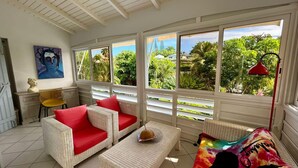 A sunlit room with wicker furniture, a painting, and a view of lush greenery.