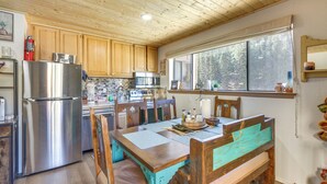 A kitchen with wooden cabinets, a stainless steel refrigerator, a dining table with chairs, and a window overlooking a forest. - Ski-In Red River Retreat: Balcony, Mountain Views! (Red River)