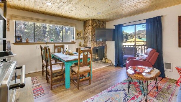 A cozy living room with a wooden ceiling, a stone fireplace, a dining area with wooden chairs, and a view of the outdoors through large windows. - Ski-In Red River Retreat: Balcony, Mountain Views! (Red River)