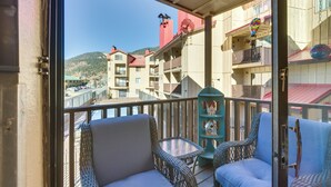 A balcony with wicker chairs and a small table, overlooking a waterfront with buildings and a mountain in the background. - Ski-In Red River Retreat: Balcony, Mountain Views! (Red River)