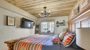 A bedroom with a wooden ceiling, a bed with a colorful blanket, a mounted TV, and a window with a view of buildings. - Ski-In Red River Retreat: Balcony, Mountain Views! (Red River)