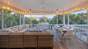 Een overdekt terras met een plafondventilator, lichtslingers en uitzicht op palmbomen en huizen.