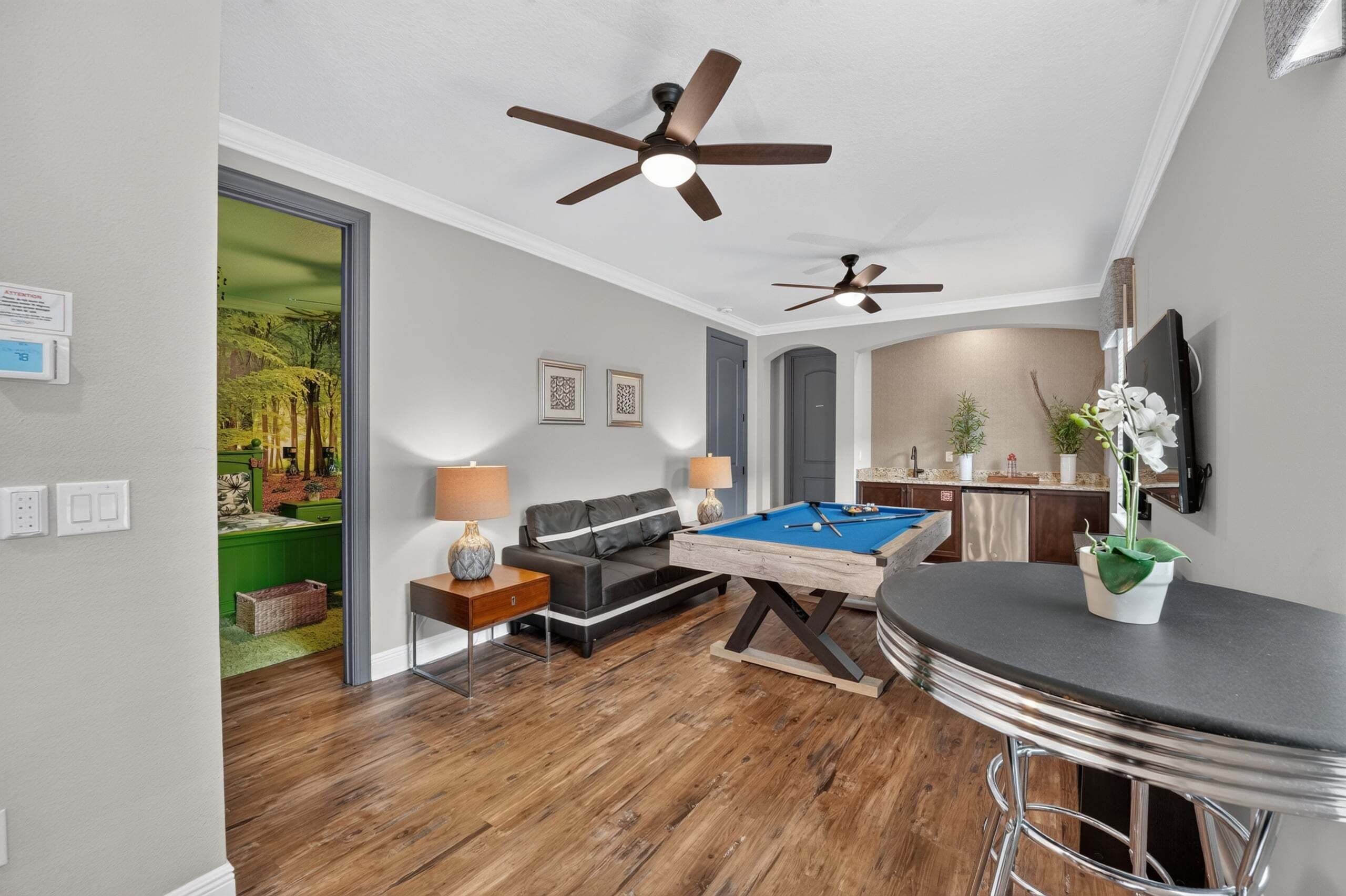 A living room with a pool table, a sofa, a ceiling fan, and a kitchen area with a TV.