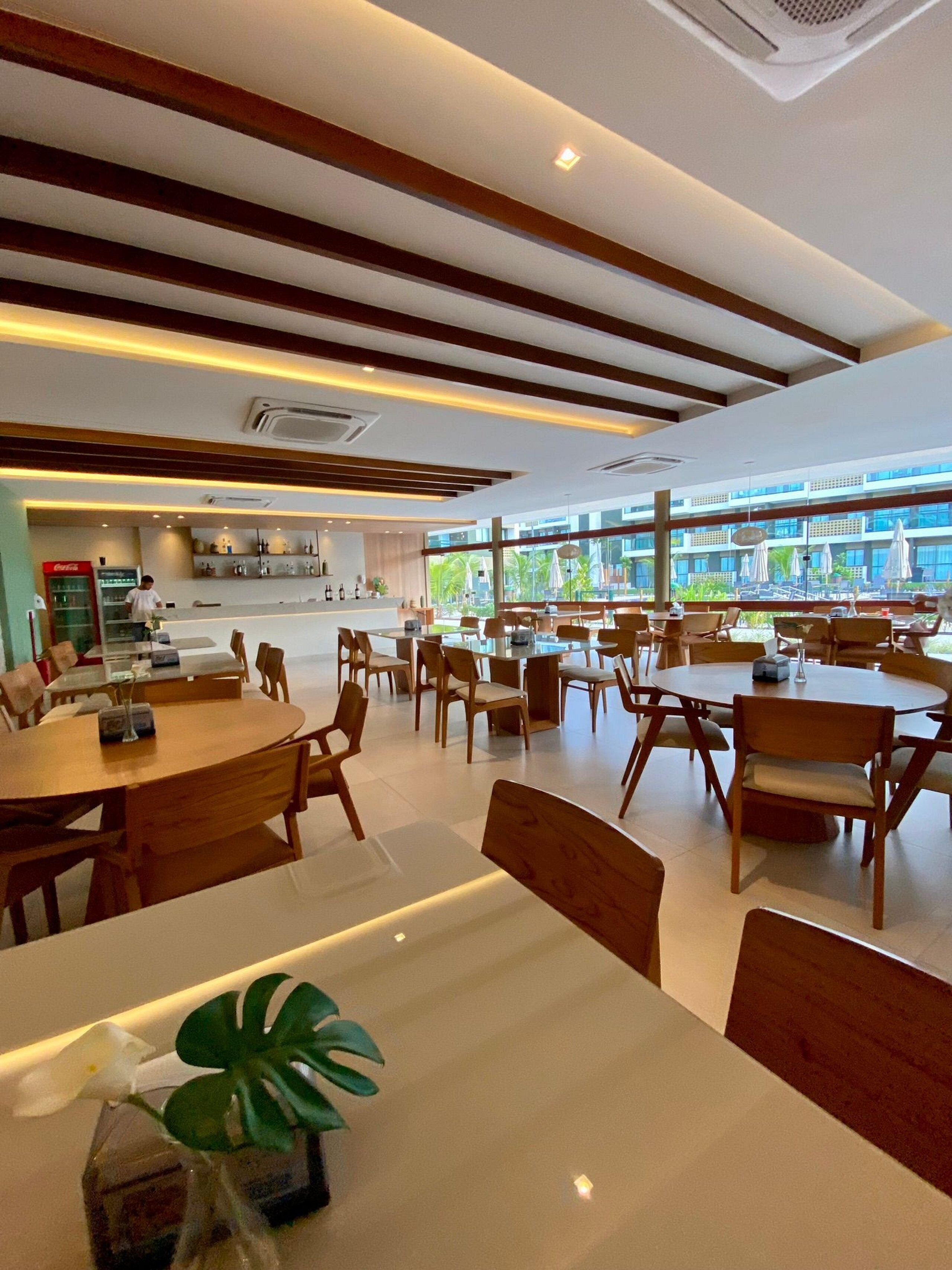 A modern dining area with wooden tables and chairs, a kitchen in the background, and large windows offering a view of the outdoors.