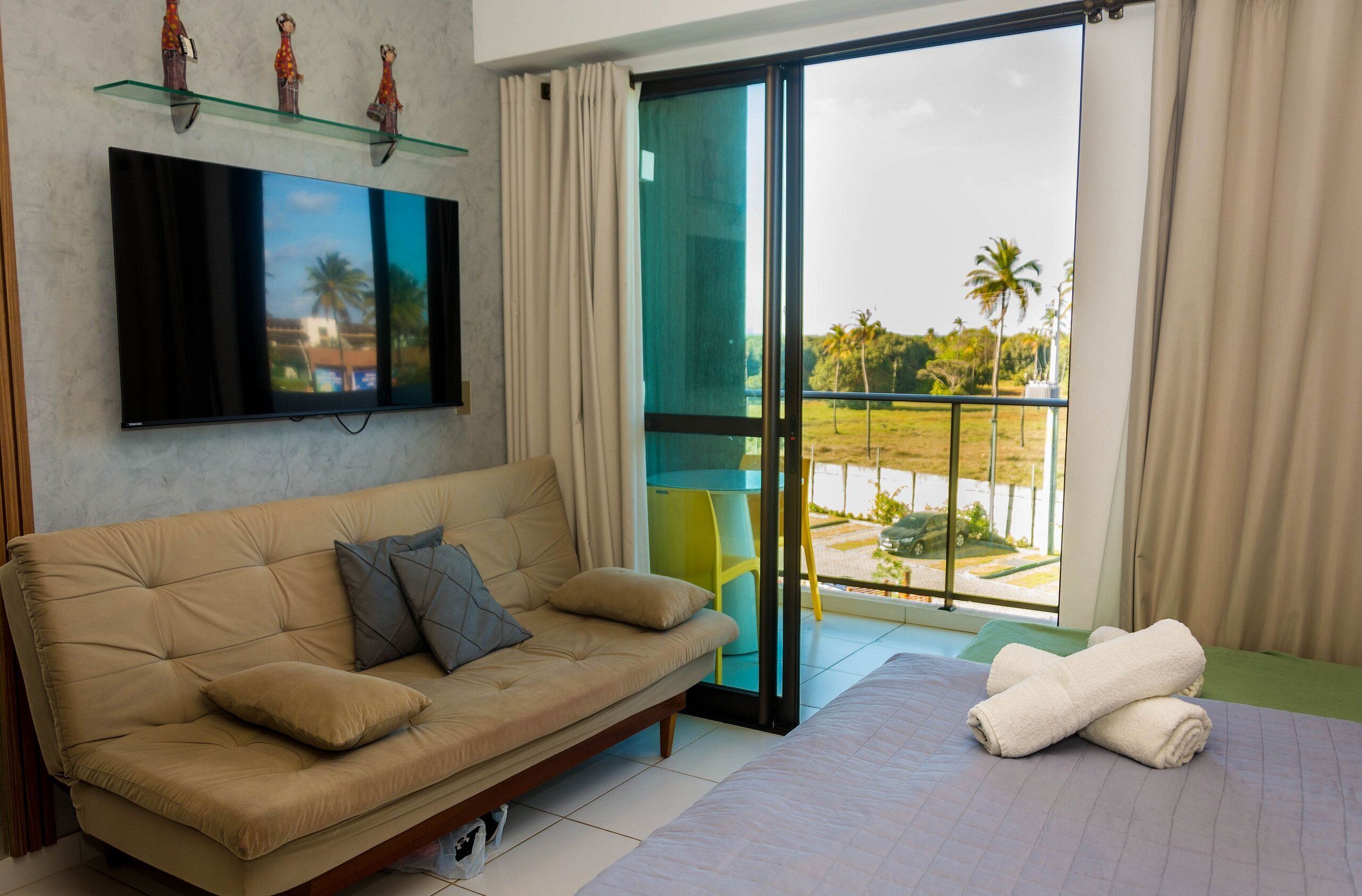 A modern hotel room with a sofa, a bed, a TV, and a balcony with a view of palm trees and a pool.